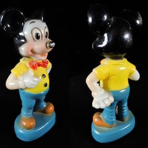 May include: A vintage Mickey Mouse figurine. The figure has a white face, black ears, and a yellow shirt with black buttons. He wears blue trousers, orange shoes, and a red bow tie. The figurine stands on a blue base.