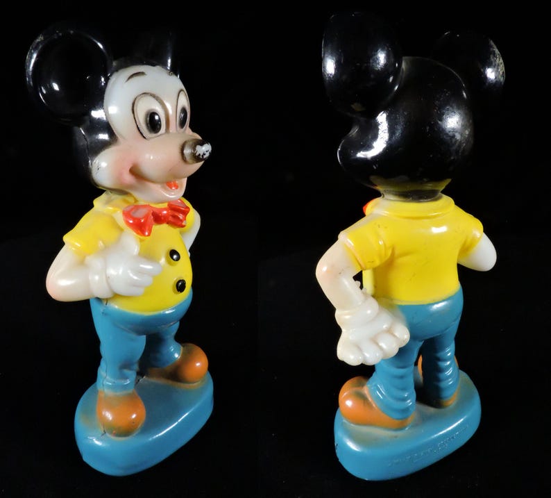 May include: A vintage Mickey Mouse figurine. The figure has a white face, black ears, and a yellow shirt with black buttons. He wears blue trousers, orange shoes, and a red bow tie. The figurine stands on a blue base.