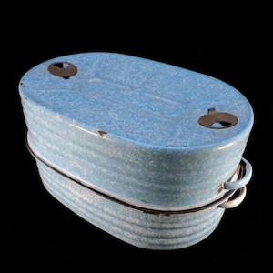 May include: A vintage, oval-shaped, blue enamel lunch pail with a speckled pattern. The lid has two metal closures and the body has two handles. The pail is trimmed with a black metal band.