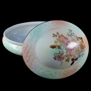 May include: An open, antique porcelain trinket box with a lid. The lid features a floral design with pink, purple, and gold accents. The box has a gold trim and a pastel color scheme of pink, green, and white.