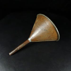 May include: A vintage copper funnel with a long, narrow spout. The funnel has a wide, conical body with a silver-colored rim. The copper has a weathered, aged appearance, suggesting it is an antique or a well-used item. The background is black.