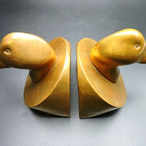 May include: A pair of gold-toned metal bookends shaped like duck heads. The bookends are on a dark surface.