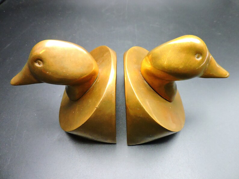 May include: A pair of gold-toned metal bookends shaped like duck heads. The bookends are on a dark surface.