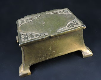 May include: A small, gold-colored metal box with a decorative, carved design on the top and four legs. The box is closed.