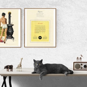 Puede incluir: Dos impresiones enmarcadas en una pared blanca, una con una ilustraci&oacute;n vintage de una mujer en traje de ba&ntilde;o blanco de pie junto a una estufa de le&ntilde;a negra, y la otra con un calendario amarillo para noviembre de 1962. Un gato gris est&aacute; acostado en una mesa de madera con una figurita de jirafa, una figurita de hipop&oacute;tamo y una radio vintage.