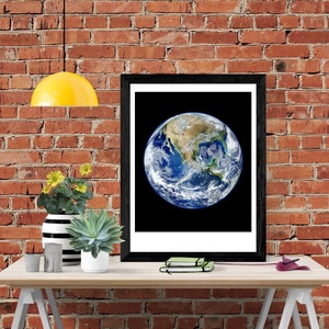 May include: A framed print of a photograph of Earth with North America visible, hanging on a brick wall above a wooden table with a yellow lamp, a vase with yellow flowers, a succulent plant, and a stack of notebooks.