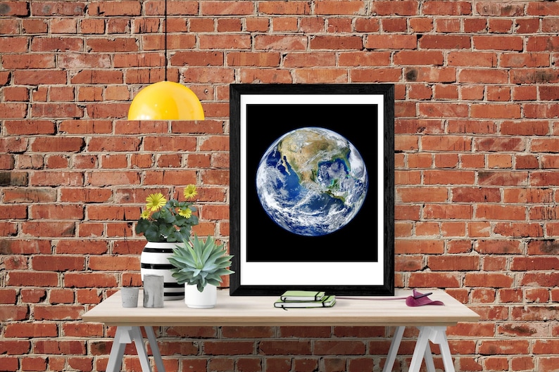 May include: A framed print of a photograph of Earth with North America visible, hanging on a brick wall above a wooden table with a yellow lamp, a vase with yellow flowers, a succulent plant, and a stack of notebooks.