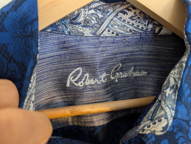 K&ouml;nnte beinhalten: Ein blaues Hemd mit Knopfleiste, das au&szlig;en ein dunkelblaues Blumenmuster und am Innenkragen sowie an der Knopfleiste einen kontrastierenden wei&szlig;-blauen Paisleydruck aufweist. Das innere Nackenetikett zeigt den gestickten Text Robert Graham.