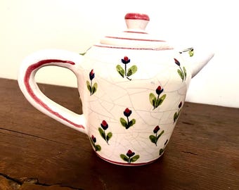 May include: A white ceramic teapot with a lid, handle, and spout. The teapot is decorated with a floral pattern of red buds and green leaves. The handle and rim are accented with red stripes. The teapot sits on a wooden surface.