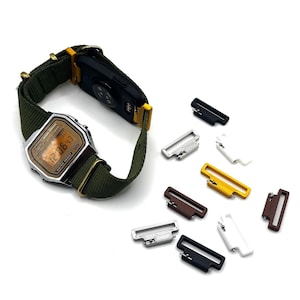 May include: A digital wristwatch with a silver-tone rectangular case, a dark olive green nylon strap, and a gold-toned digital display face. The watch is accompanied by several small, rectangular watch strap adapters in various colors.