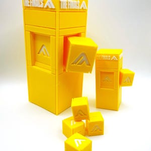 以下が含まれることがあります： 鮮やかな黄色の3Dプリントされたゲームピースのセット。メインの構造は長方形で、「THE FINALS」の文字が入っています。白い「A」のロゴが付いた小さな立方体がいくつか、構造の周りに散らばっています。