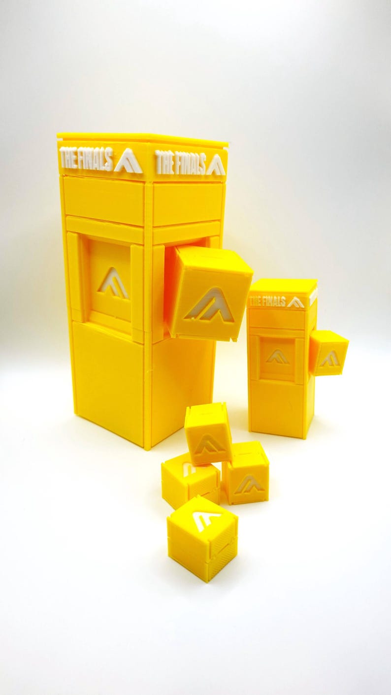 以下が含まれることがあります： 鮮やかな黄色の3Dプリントされたゲームピースのセット。メインの構造は長方形で、「THE FINALS」の文字が入っています。白い「A」のロゴが付いた小さな立方体がいくつか、構造の周りに散らばっています。