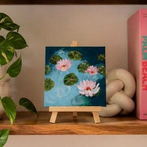May include: A square canvas painting on a small wooden easel depicting three light pink water lilies floating on a blue water surface with green lily pads. The painting features visible brushstrokes and a vibrant color palette of blue, pink, and green.