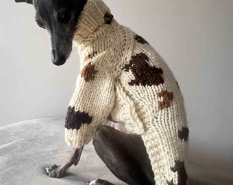 May include: A black and gray dog wearing a cream-colored, chunky knit sweater with brown spots. The sweater has a high collar and covers the dog's front legs and torso. The dog is sitting on a gray surface.