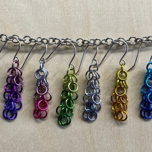 May include: Six pairs of dangle earrings with a chainmail design. The earrings are in a rainbow of colors, including purple, pink, green, silver, gold, and blue.