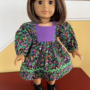 May include: A doll wearing a floral print dress with a purple bodice and green trim. The dress has long sleeves and is paired with black lace-up boots. The doll has brown hair and is standing on a red surface.