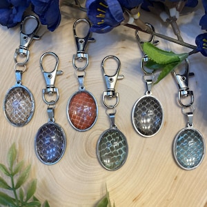 Puede incluir: Una colección de llaveros con colgantes ovalados con varios colores y patrones. Los colgantes están engastados en marcos de metal plateado y sujetos a cierres de mosquetón plateados. Los patrones se asemejan a piel de serpiente en tonos marrón, naranja, verde y azul.