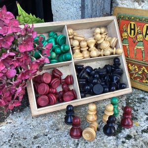 Puede incluir: Una caja de madera con compartimentos llenos de piezas de juego verdes, negras y crema. La caja está abierta y las piezas son visibles. La caja está sentada en una mesa con un patrón floral.
