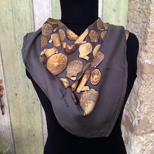 May include: A gray silk scarf featuring a pattern of seashells in shades of brown and tan. The scarf is draped over a black turtleneck. The text "HERMES-PARIS" is visible on the scarf.
