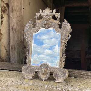 Può includere: Uno specchio in metallo dorato ornato da una cornice decorativa. Lo specchio è rettangolare e ha un bordo bisellato. La cornice è molto dettagliata con intagli intricati e volute. Lo specchio riflette un cielo nuvoloso.