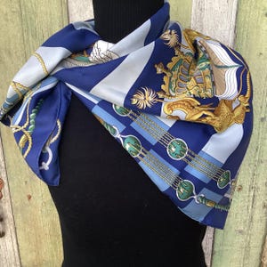 May include: A silk scarf in shades of blue, light blue, and gold. The scarf features nautical and heraldic designs, including ropes, shields, and stylized figures. The scarf is draped over a black form.