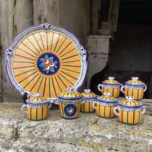 Op de afbeelding: Een set van negen keramische serveerschalen met gele, blauwe en groene bloemmotieven. De schalen zijn in een cirkel gerangschikt rond een grotere, gele en blauwe keramische schotel met een bloemmotief.