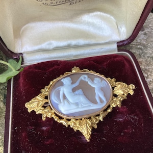 Puede incluir: Broche antiguo dorado con un camafeo ovalado que representa figuras clásicas. El camafeo está engastado en un marco de filigrana ornamentado. El broche se exhibe sobre una superficie de terciopelo burdeos dentro de un joyero vintage.