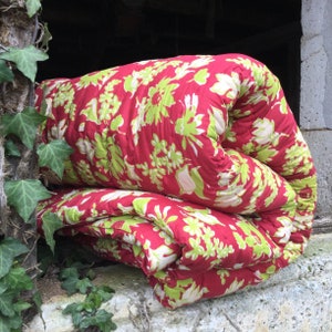 Op de afbeelding: Een opgerolde futonmatras met een rood en groen bloemenpatroon ligt op een stenen oppervlak.