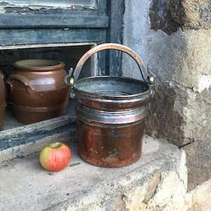 May include: A copper bucket with a brass handle and a worn patina. The bucket is sitting on a stone surface next to a brown clay pot.