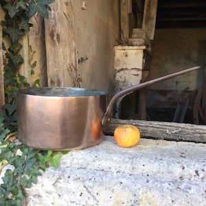 Puede incluir: Una olla de cobre con un mango largo se encuentra sobre una superficie de piedra junto a una manzana amarilla. La olla está hecha de cobre y tiene un acabado brillante. La manzana es de color amarillo brillante y tiene una piel lisa.