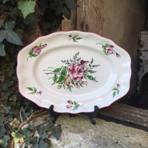 May include: A white oval serving platter with a pink rim and a floral design featuring a large pink rose with green leaves and yellow accents.