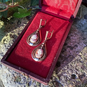 May include: A pair of vintage cameo earrings presented in an open, red velvet-lined jewellery box. The earrings have a teardrop shape, with a cameo portrait set in a silver-tone frame. The box's interior provides a striking contrast to the earrings.
