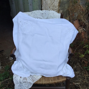 May include: White embroidered tablecloth with a delicate floral pattern, draped over a wooden chair with a woven seat.