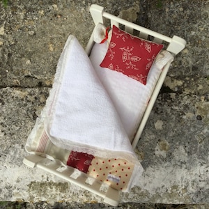 May include: A white doll bed with a red and white patterned quilt and a red pillow with white floral designs. The bed is made of wood and has a white finish.
