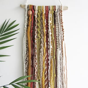 May include: A handcrafted wall hanging with a mix of textured yarns in brown, yellow, and white hues. The yarns are secured to a natural wooden dowel and adorned with wooden beads. The wall hanging is suspended by a white string.