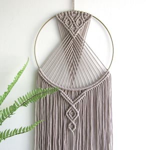 May include: A beige macrame wall hanging with a gold metal hoop and a geometric pattern. The hanging has long fringe strands.