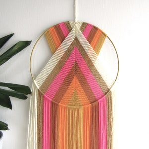 May include: A colourful macrame wall hanging with a gold hoop and white, pink, orange, and brown yarn. The yarn is woven into a geometric pattern with fringe hanging from the bottom.