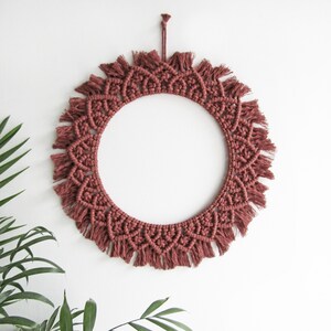 May include: A circular macrame wall hanging in a muted red-brown hue. The handcrafted piece showcases detailed knotwork and a fringed edge. It is suspended by a knotted cord, offering a stylish, bohemian accent for interior spaces.