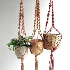 May include: Three macramé plant hangers in shades of brown. One holds a white ceramic pot with greenery, the others have woven baskets. Each hanger has a knotted design with a tassel at the bottom, creating a bohemian aesthetic.