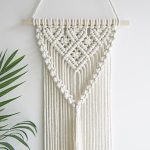 May include: Cream-coloured macrame wall hanging. The top is attached to a natural wooden dowel. The design features a geometric pattern at the top, transitioning into a triangular shape with long, vertical fringe.