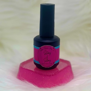 May include: A dark blue nail polish bottle with a pink and teal label that reads "Stay & Slay" sits on a pink, glittery, hexagonal base. The bottle has a dark blue cap. The background is a soft, white, fluffy texture.