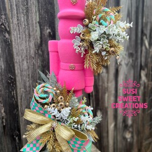 May include: A pink, flocked nutcracker figurine with gold and teal accents. The nutcracker is decorated with a bow, candy canes, and greenery. The nutcracker is sitting on a teal box with the text "Susi Sweet Creations".