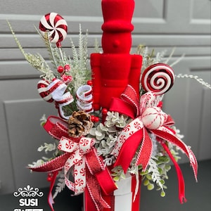 May include: A festive Christmas centerpiece featuring a red nutcracker, peppermint candies, and decorative bows. The arrangement is in a red and white striped container, with greenery and berries. The text "SUSI SWEET CREATIONS" is visible.