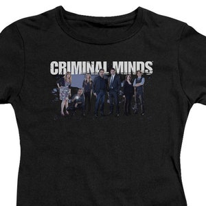 May include: Black t-shirt with the text "CRIMINAL MINDS" in white letters. The image features a group of people standing in front of a dark background.