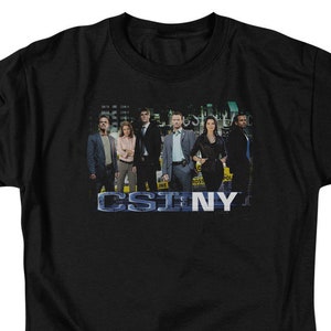 May include: Black t-shirt with a group of people in a line wearing suits and holding evidence bags. The text "CSI NY" is printed on the shirt.