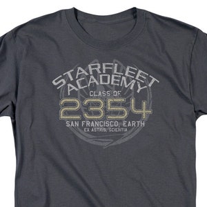 May include: A dark gray t-shirt with a vintage-style graphic of the Starfleet Academy logo. The logo features the text "Starfleet Academy" in a circle with the text "Class of 2354" below it. The text "San Francisco, Earth" and "Ex Astris, Scientia" are also included in the logo.