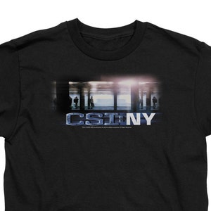 May include: Black t-shirt featuring the text "CSI NY" in a blue and white design. The graphic includes a blurred image of a city scene. The shirt is made of a soft material.