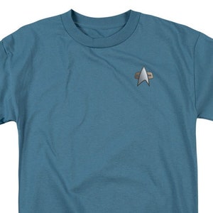 May include: A blue short-sleeved t-shirt with a silver Starfleet insignia on the left chest.