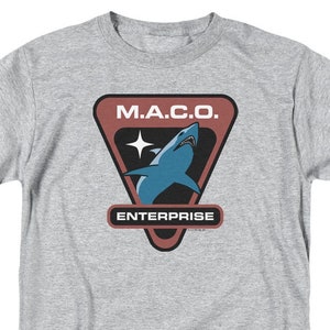 Peut inclure: T-shirt gris clair avec un motif. Le motif présente le texte "M.A.C.O." au-dessus d'une illustration de requin bleu et le mot "ENTERPRISE" en dessous. Le motif est encadré dans un triangle marron et noir.