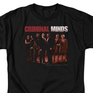 May include: Black t-shirt with a red and white graphic of the text "CRIMINAL MINDS" and a group of people standing in front of a blurred background.